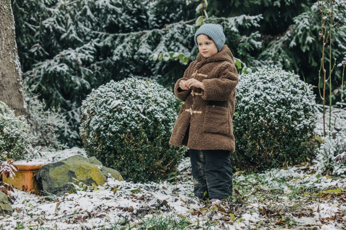 Kleines Kind mit Wintermütze und Mantel in einem verschneiten Garten