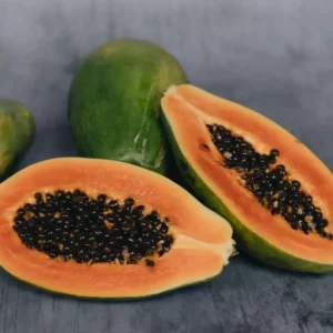 Zwei Papayahälften mit orangefarbenem Fruchtfleisch und schwarzen Kernen, dahinter eine ganze grüne Frucht auf grauem Untergrund